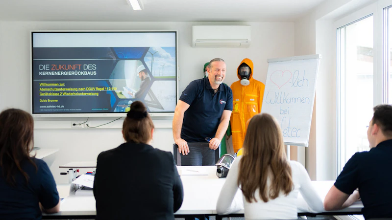 lehrer vor einer tafle mit einer blau-grauen Grafik. Schüler und Schülerinnen sitzen davor.