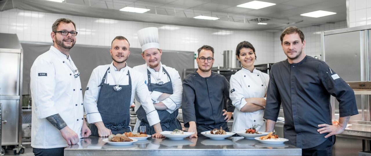 Männer und Frauen in typischer Kochbekleidung in weiß und grau in einer Küche mit Edelstahlschränken und hinter einer Theke stehend.