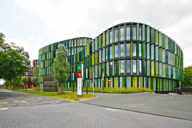 Wellenförmiges mehrstöckiges Gebäude mit viel Glas und grünen und gelben Fensterelementen, das an einer gepflasterten Straße steht.