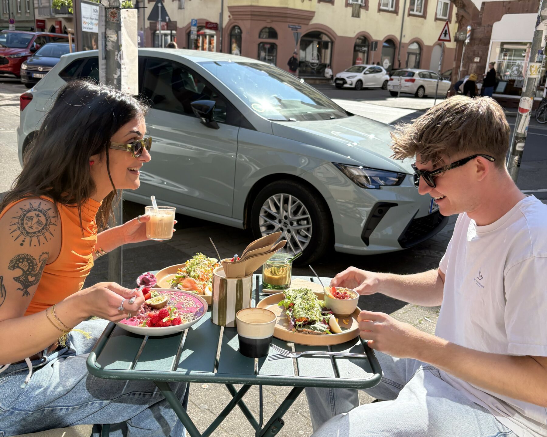 Zwei junge Menschen sitzen an einem kleinen Tisch in der Sonne und frühstücken. Im Hintergrund parkt ein Auto, die Sonne spiegelt sich in der Windschutzscheibe.