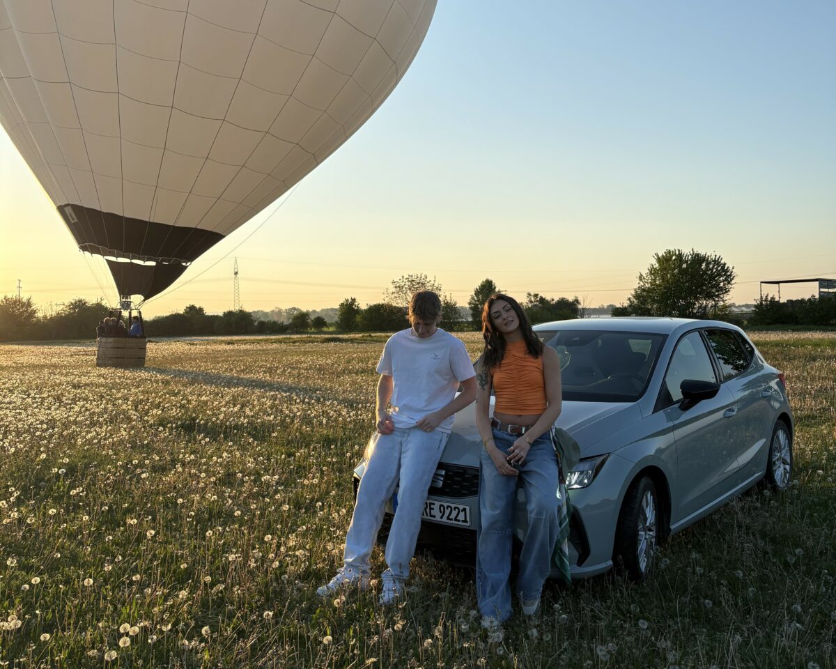 Ein junger Mann und eine junge Frau lehnen nebeneinander an der Motorhaube eines weißen Autos. Das Auto steht auf einer Wiese, im Hintergrund sieht man einen Heißluftballon, Bäume und den Abendhimmel.