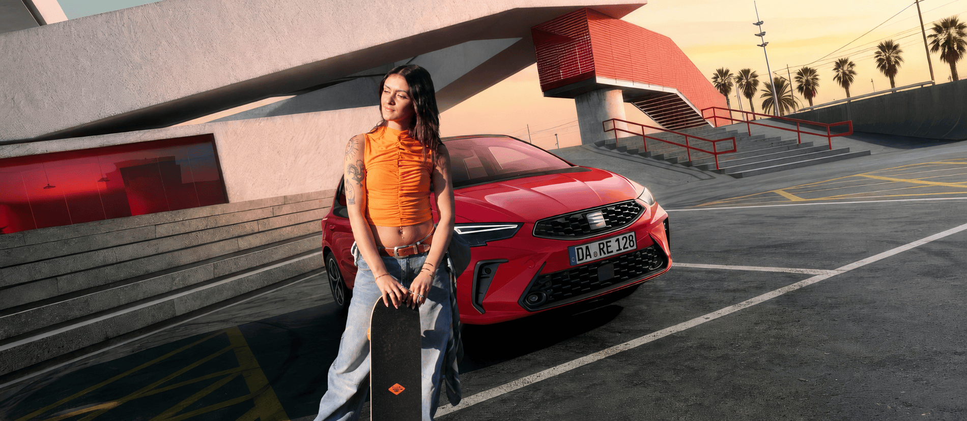 bigFM-Moderatorin Filiz in Jeans und einem orangenen Top steht vor einem roten SEAT Ibiza. In den Händen hält sie ein Skateboard, im Hintergrund sieht man ein Gebäude mit einem Geländer und Palmen vor abendlichem Himmel.