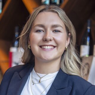 Blonde, lächelnde junge Frau mit weißem Shirt und dunkelblauem Blazer vor einem Weinregal.